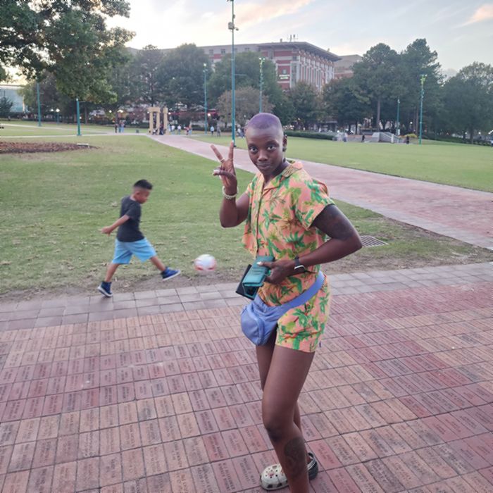 Imani throwing a peace sign in a park at sunset while a child plays soccer behind her.