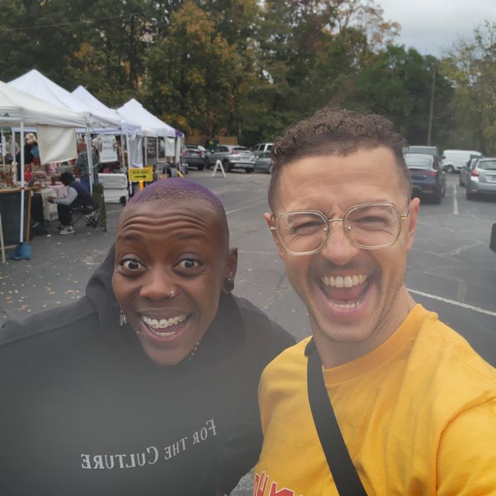 Imani and Julian grinning in a selfie at an outdoor market, Imani in a &lsquo;For the Culture&rsquo; hoodie.