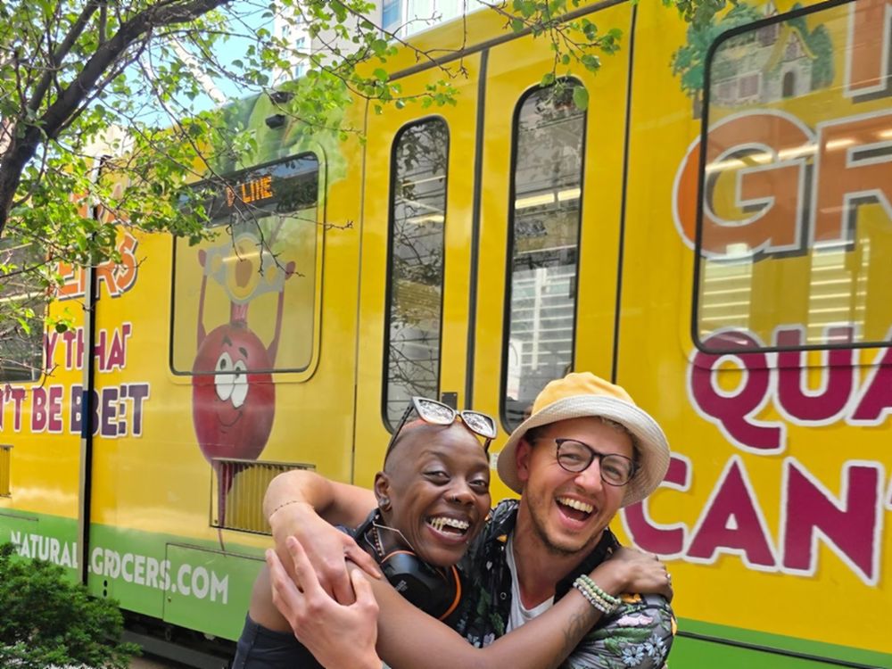 Imani and Julian embracing and laughing in front of a bright yellow city tram.