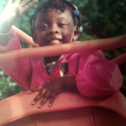 Imani Turnbull-Brown as a young child, grinning on a swing in dappled sunlight.